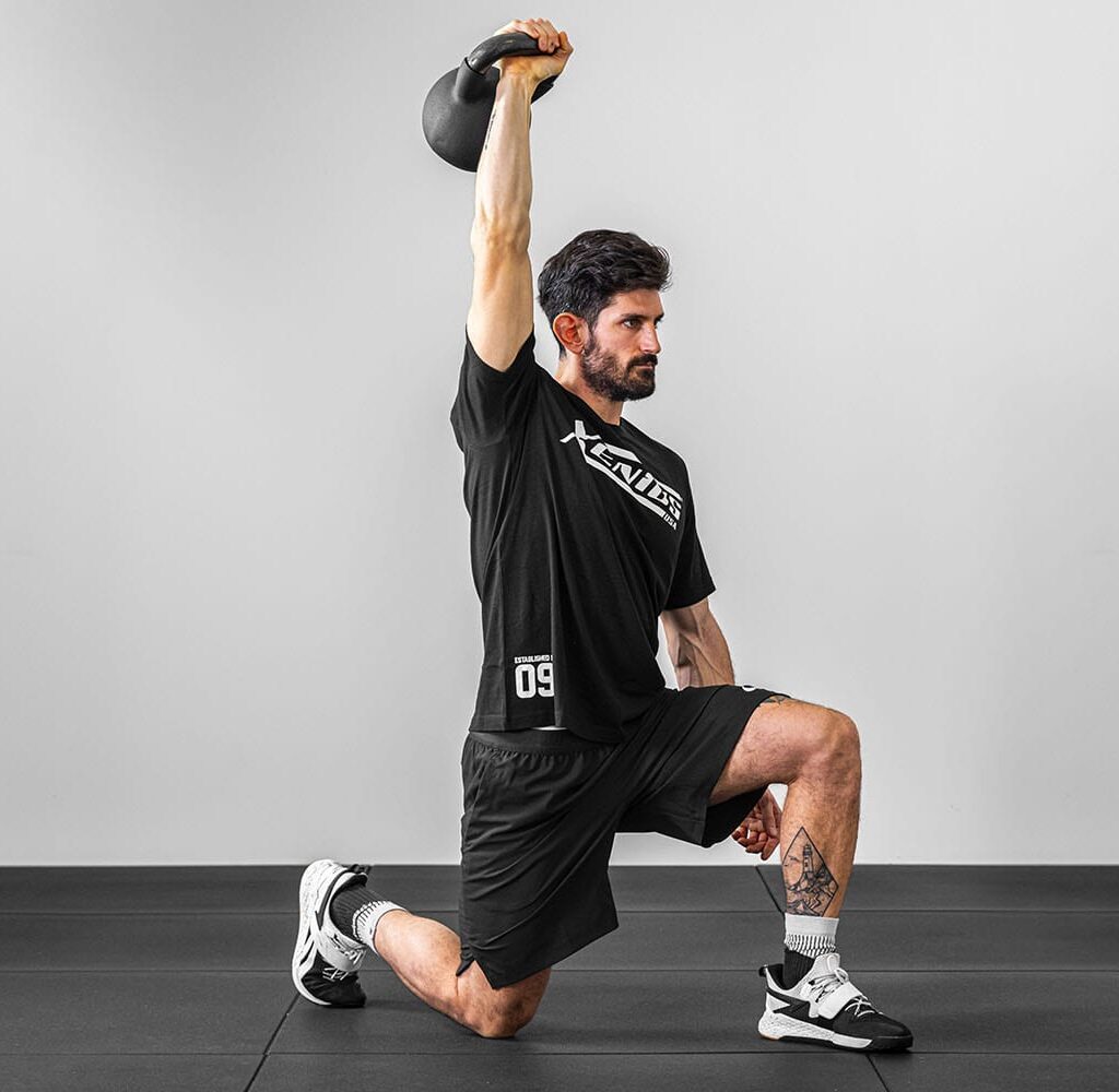 Un joven realizando un paso hacia adelante con una kettlebell que sostiene con el brazo extendido sobre su cabeza.