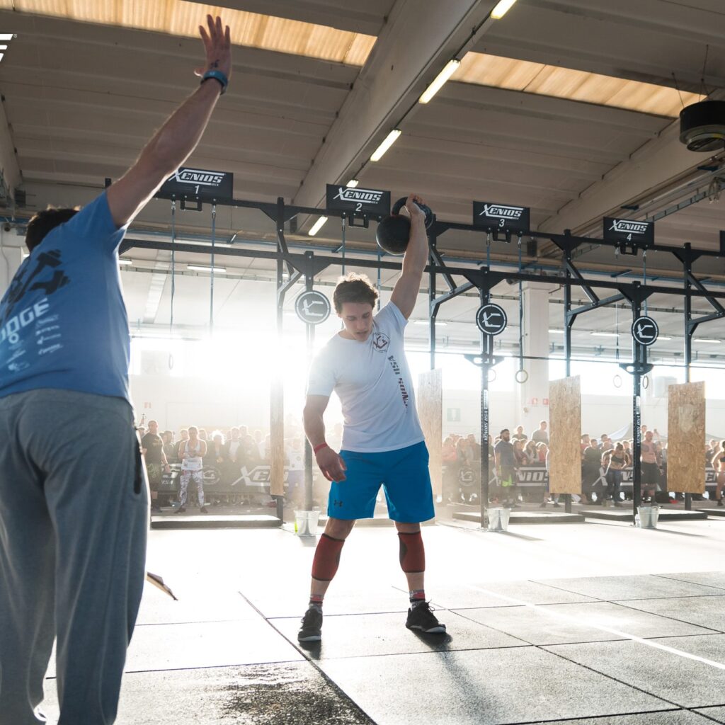 Un joven realiza un Clean & Press con kettlebells durante una competición. El área de entrenamiento está despejada y se pueden ver las siluetas de los espectadores en el fondo, observando. Un juez evalúa la ejecución del competidor desde atrás.