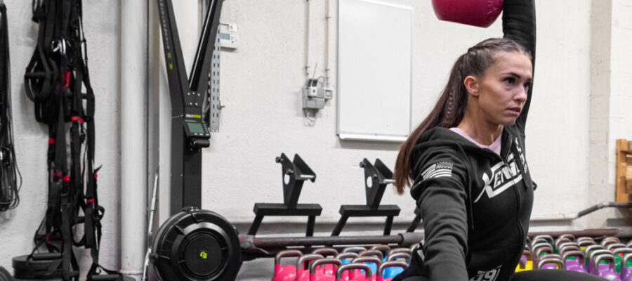 Una chica realiza un entrenamiento de kettlebell en un gimnasio, realizando una sentadilla profunda con una kettlebell levantada con un brazo sobre su cabeza. Detrás, en el suelo cerca de la pared, hay numerosas kettlebells ordenadas por color. También se ven otros equipos de entrenamiento apoyados en la pared.