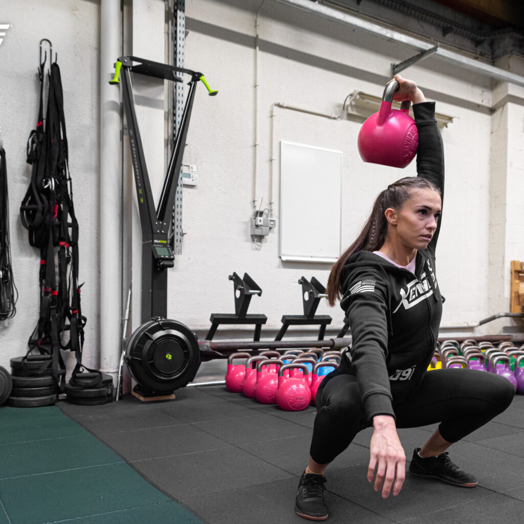 Una chica realiza un entrenamiento de kettlebell en un gimnasio, realizando una sentadilla profunda con una kettlebell levantada con un brazo sobre su cabeza. Detrás, en el suelo cerca de la pared, hay numerosas kettlebells ordenadas por color. También se ven otros equipos de entrenamiento apoyados en la pared.